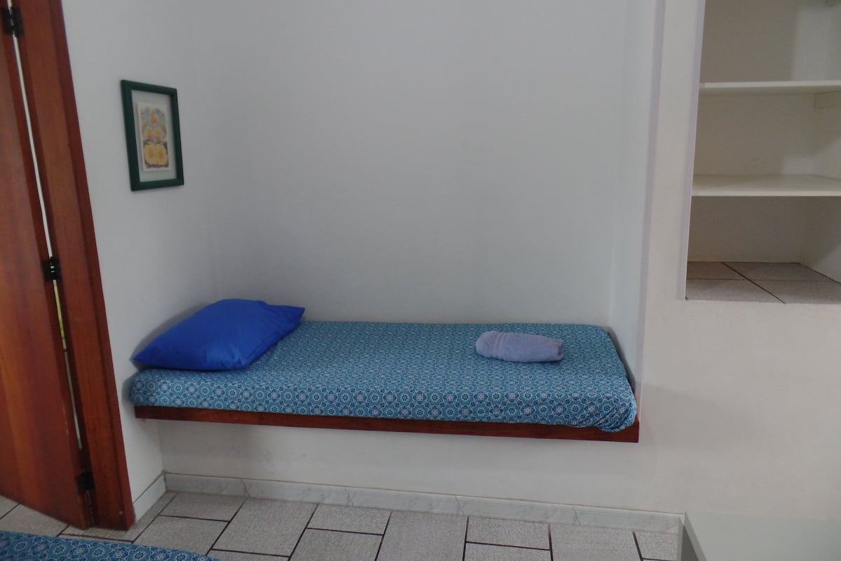 Ein kleiner, schmaler Raum mit einer Bank als Bett, einem blauen Kopfkissen und einer kleinen Regalzeile.