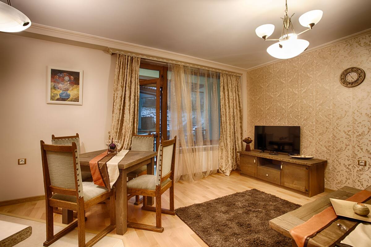 A dining area with a wooden table and chairs, a television on a wooden cabinet, and a patterned wallpaper wall.