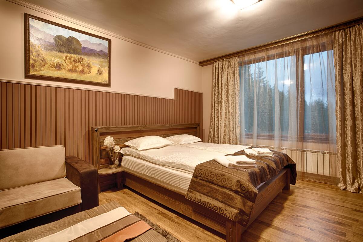 A bedroom with a wooden bed, a nightstand with a vase of flowers, a wooden bench, and a painting on the wall.