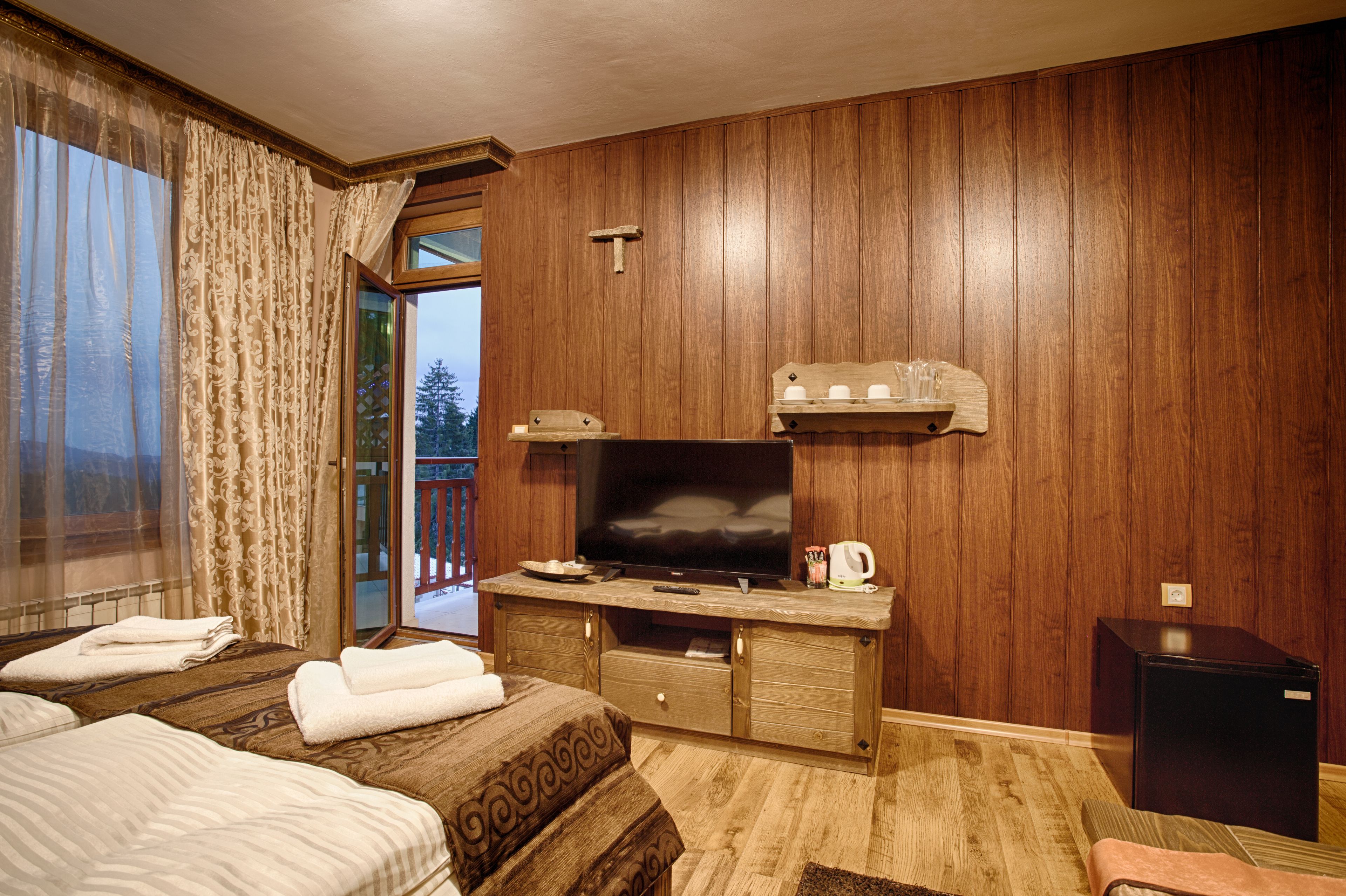 A hotel room with wooden panel walls, a television, a small refrigerator, and a sliding glass door leading to a balcony.