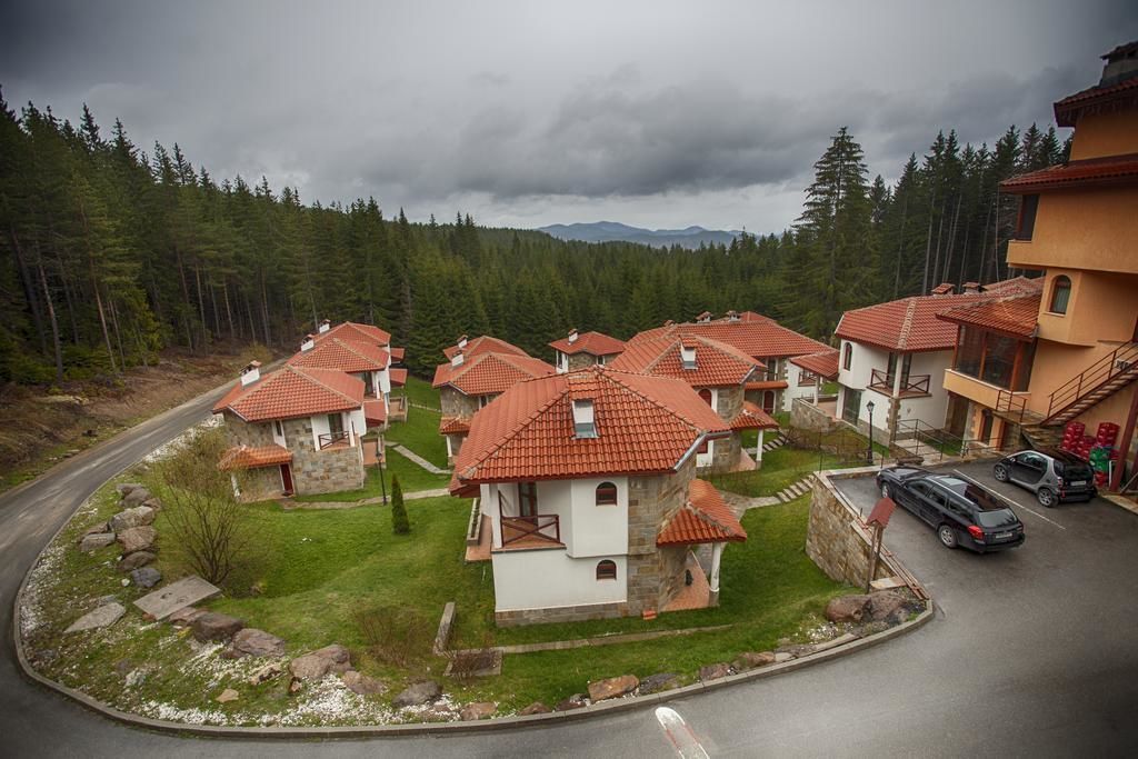 A cluster of stone houses with red-tiled roofs, arranged in a semi-circular pattern around a central road, set against a backdrop of dense forest and mountains.