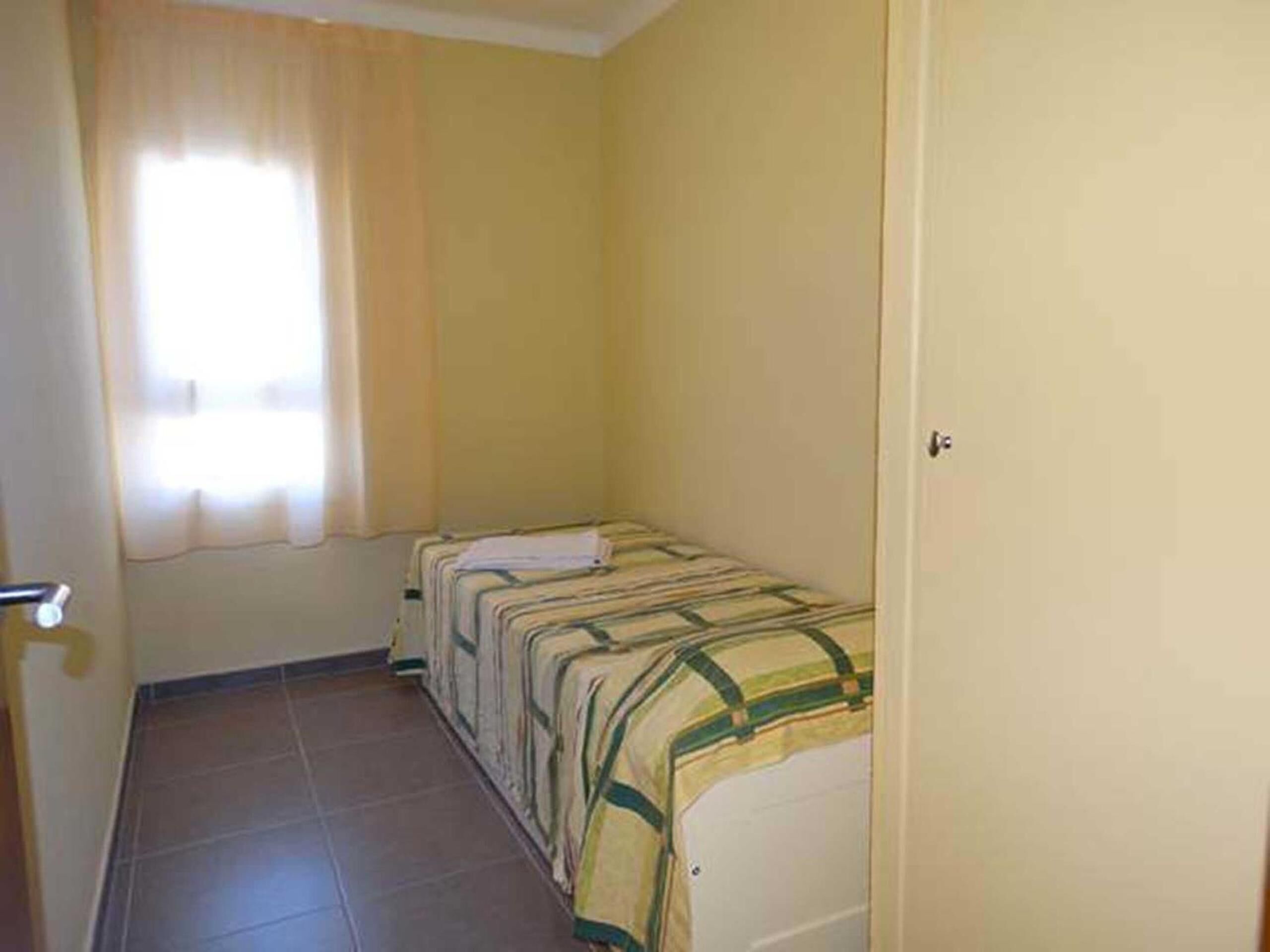Una habitación pequeña con cama individual, una pared amarilla sencilla, una ventana con cortinas y suelo de baldosas.