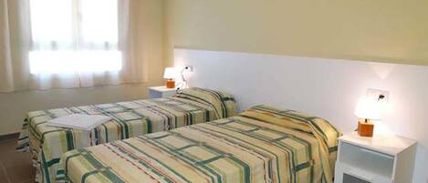 Una habitación de hotel con dos camas, una mesita de noche con lámpara y una ventana con cortinas.