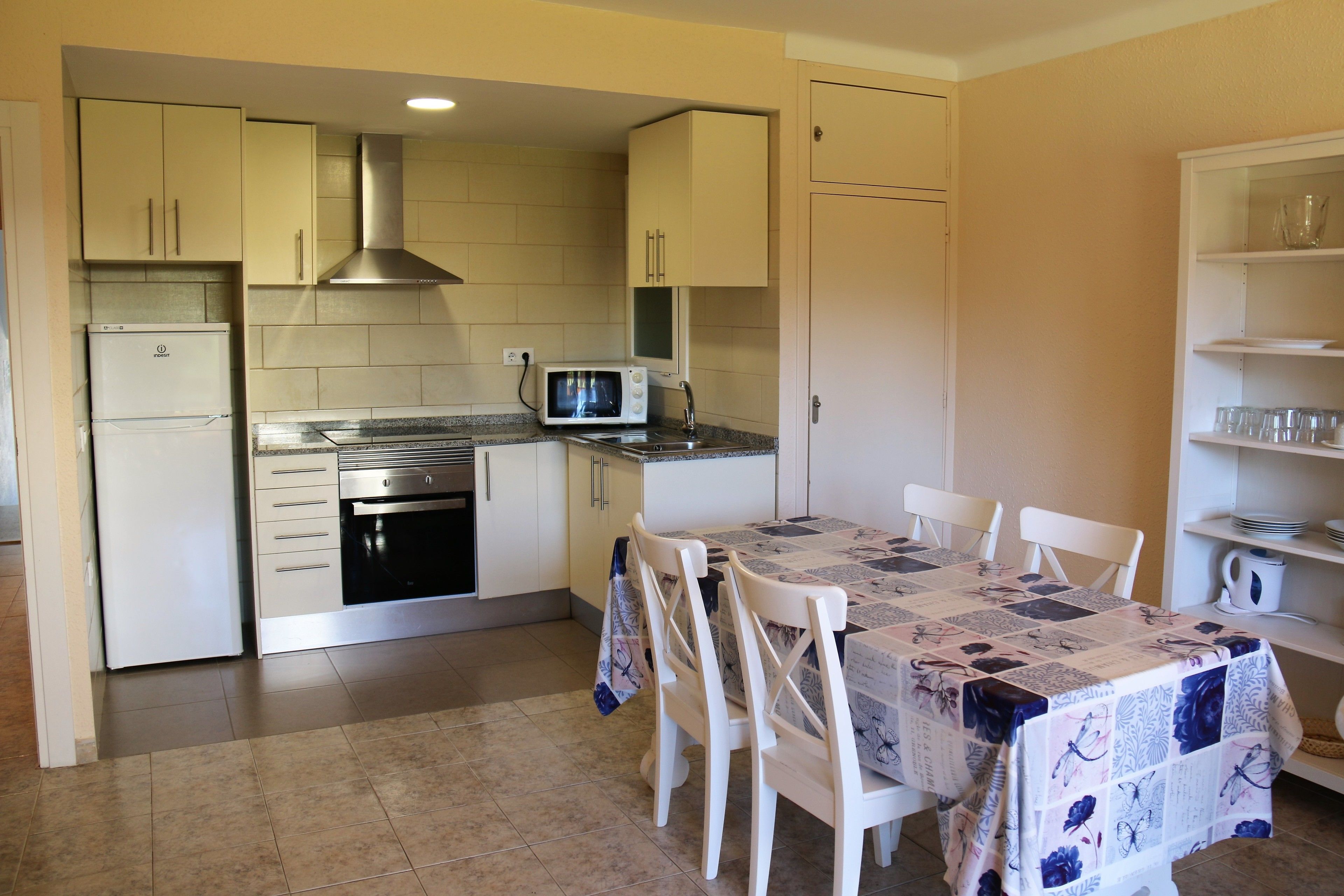 A compact kitchen with a refrigerator, microwave, stove, and sink. There is a dining table with chairs and a tablecloth.