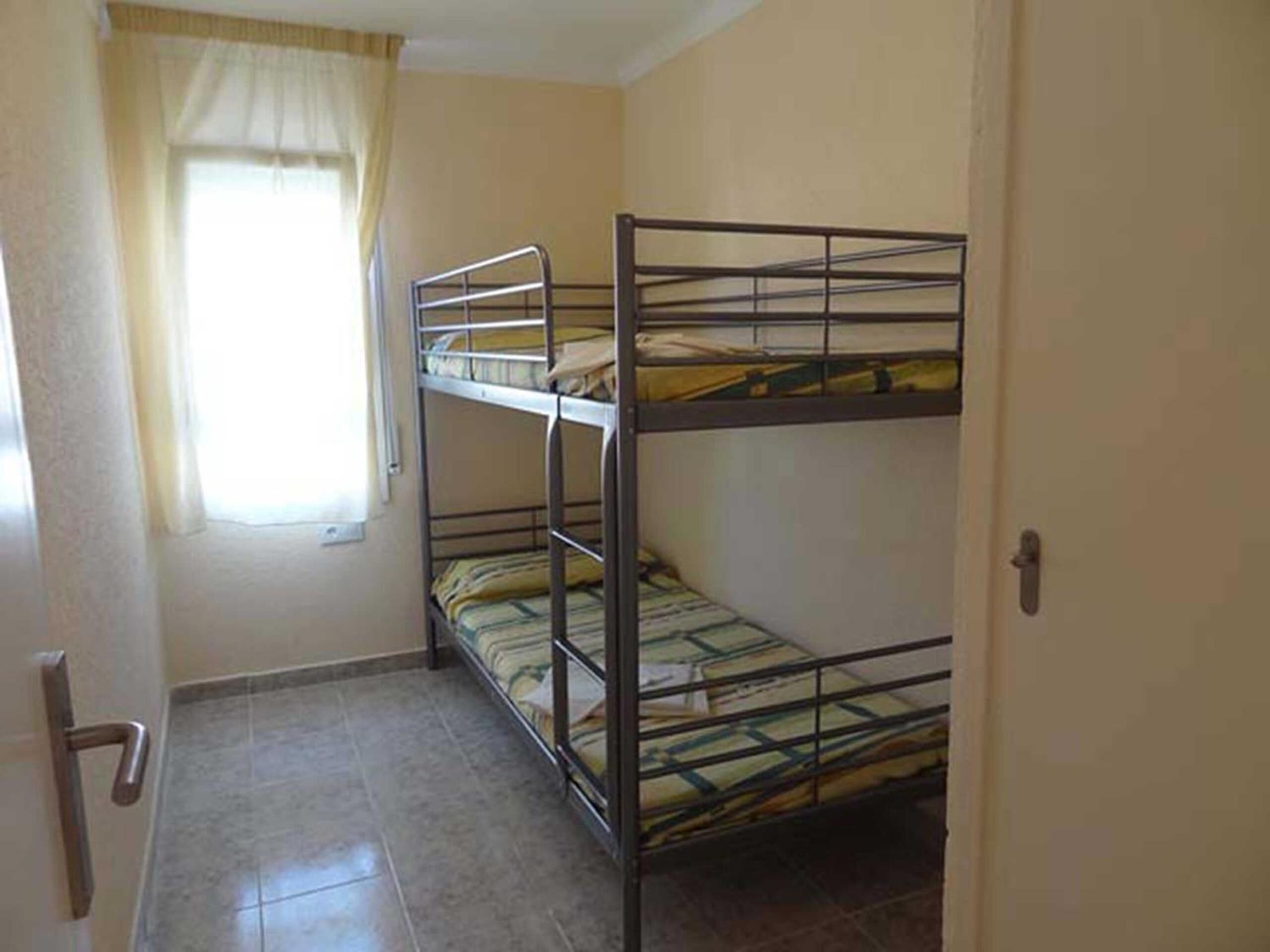 Una habitación pequeña con camas literas, ventana con cortinas y piso de baldosas.