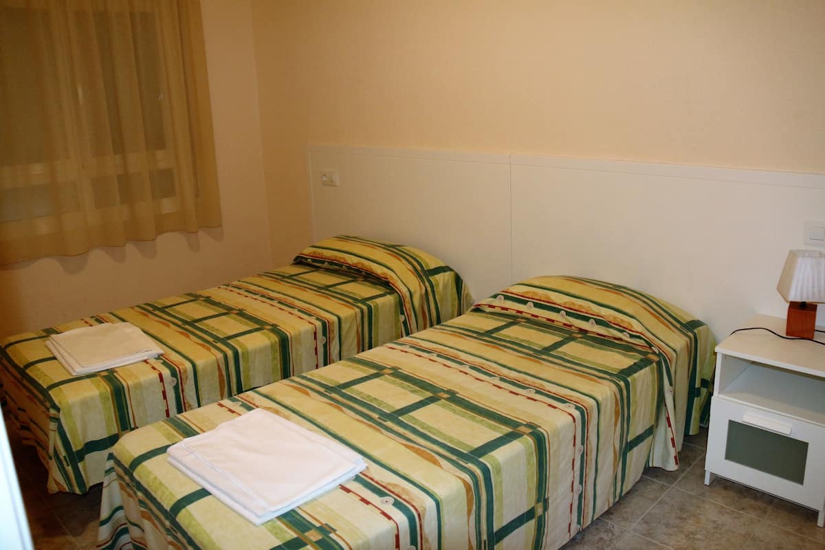 Ein Hotelzimmer mit zwei Einzelbetten, beide mit einer gestreiften Bettdecke und einem zusammengeklappten weißen Handtuch. Es gibt einen kleinen weißen Nachttisch mit einer Lampe darauf.