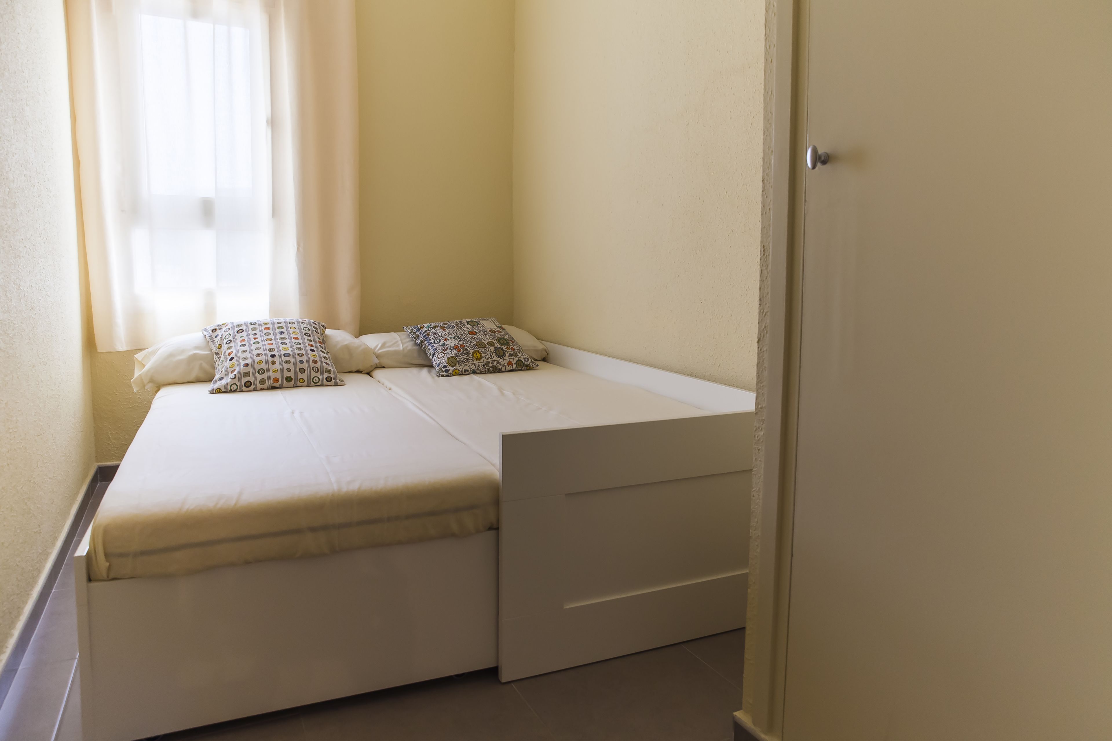 A small, single-bed room with a white mattress, two pillows, and a small storage drawer.