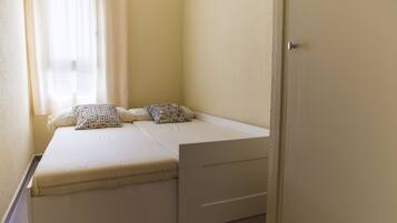A small, single-bed room with a white mattress, two pillows, and a small storage drawer.