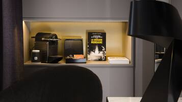 A hotel room with a seating area featuring a black chair and a white table. A shelf displays a coffee machine, a box, and a book titled "LA BRIGADE MONDAINE" by Claude Cancres.