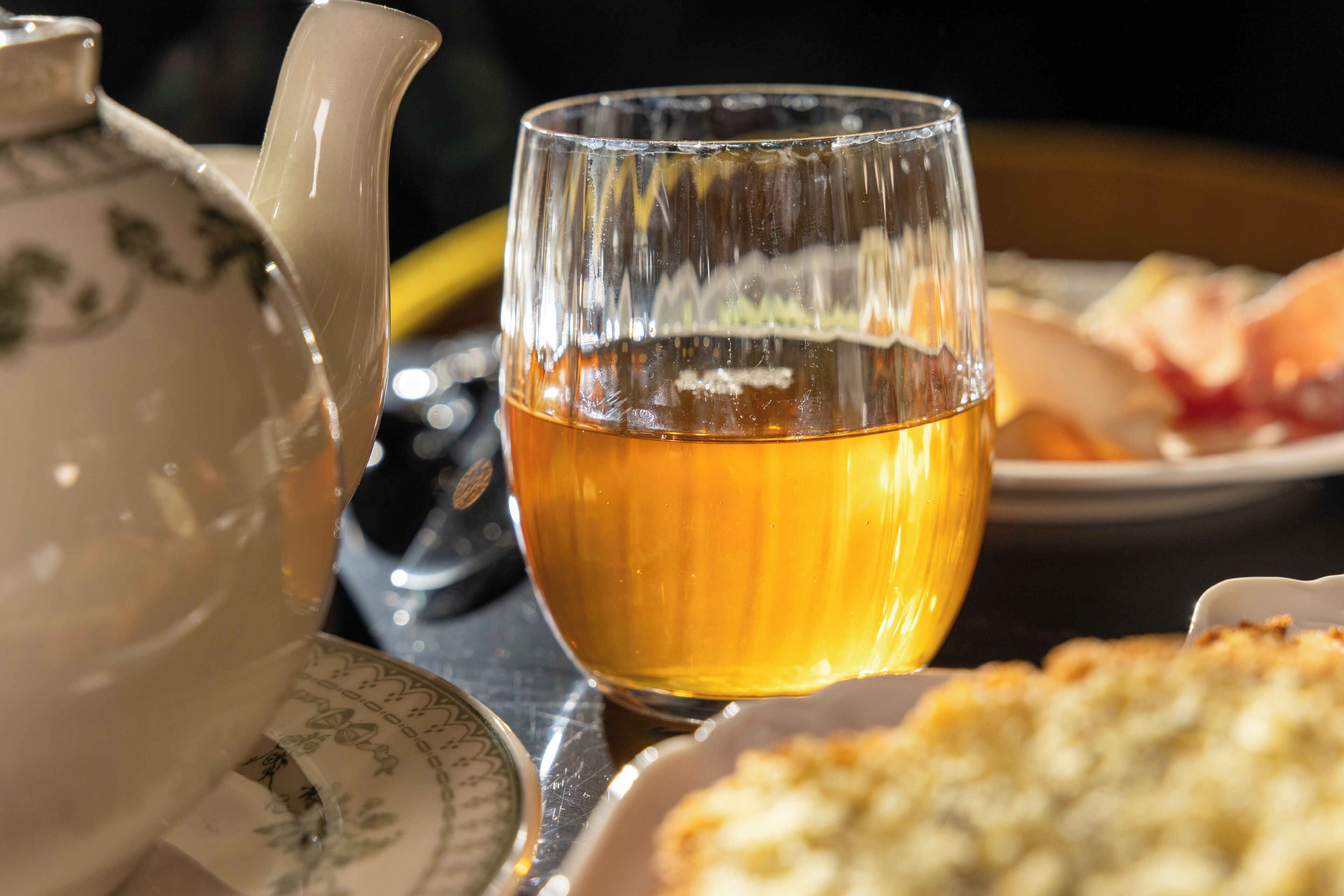 A teapot, a glass with tea, and a plate with food on a table.