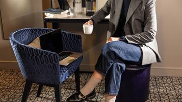 A person seated on a blue patterned chair with a laptop on a table, a cup in hand, and a desk with a bottle and books in the background.