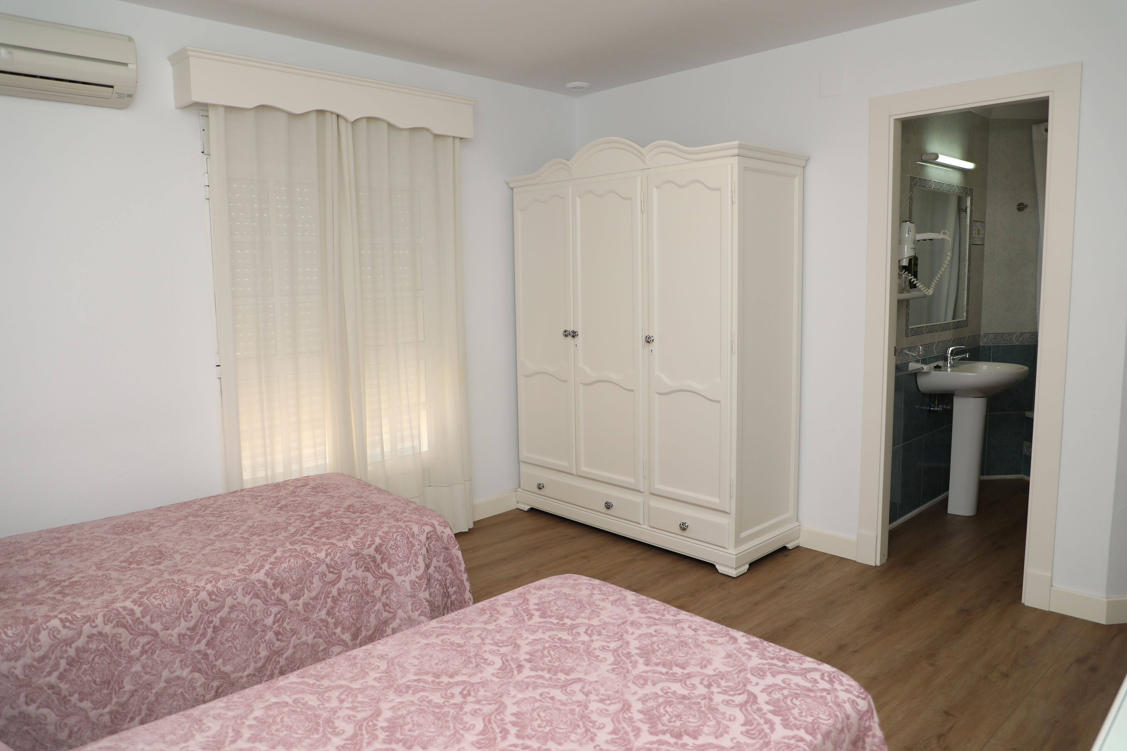 A bedroom with two beds, a wardrobe, a bathroom with a sink, and a window with curtains.