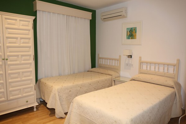 A room with two single beds, a wardrobe, a window with curtains, an air conditioning unit, and a wall-mounted lamp. - Hostal La Janda (Vejer de la Frontera)