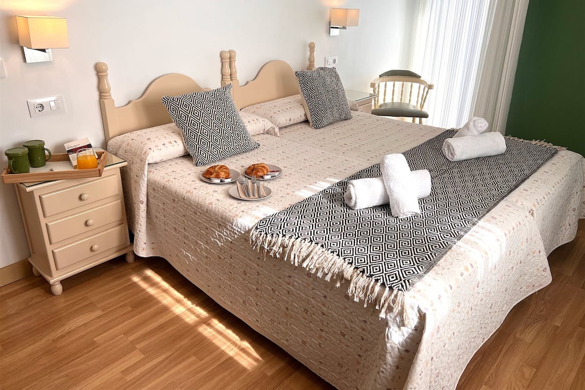 A neatly made bed with a patterned bedspread and pillows, a bedside table with a tray of croissants and a glass of orange juice, and two wall-mounted lamps.