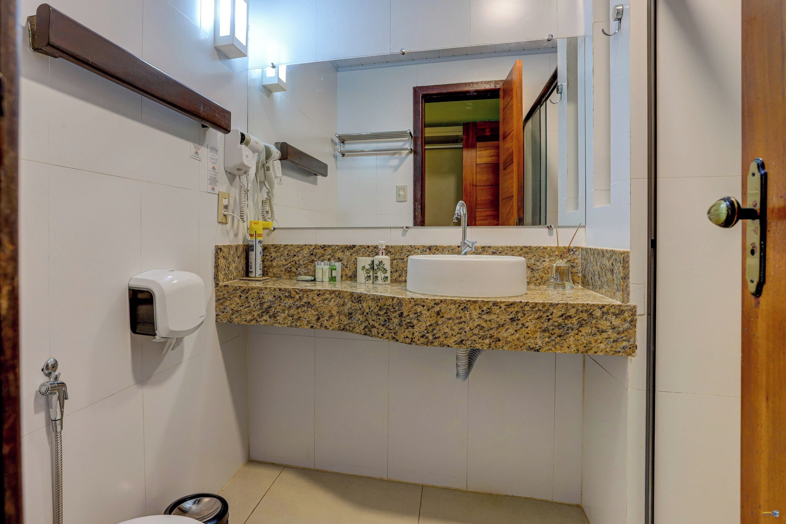A bathroom with a granite countertop sink, a handheld showerhead, a towel dispenser, and a mirror with a wooden frame.