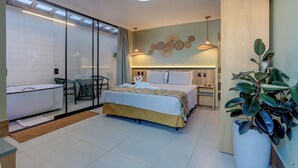 A modern hotel room with a glass partition, a bed with a floral bedspread, a bedside table, a potted plant, and a wall-mounted light fixture. - Pousada Praia Bela (Ilhéus)