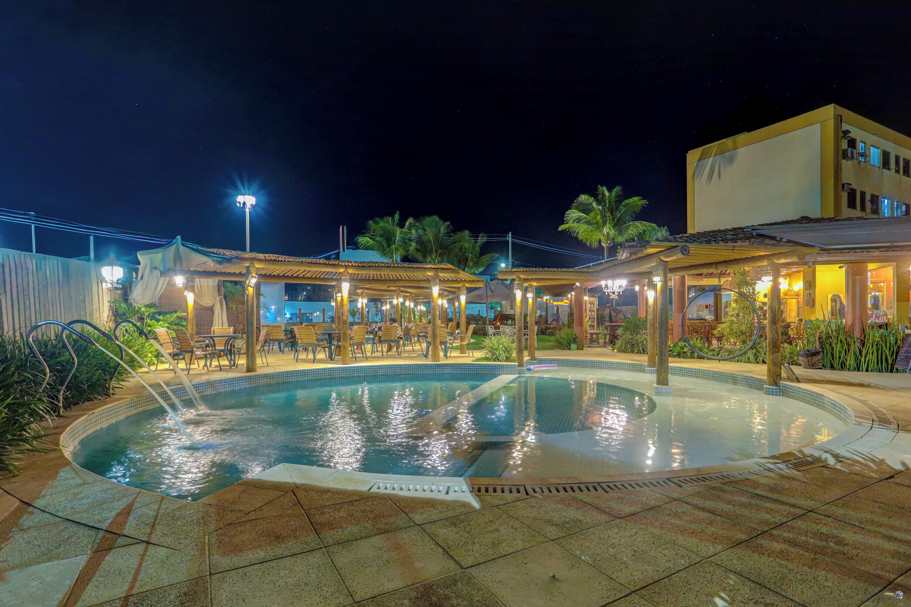 Une piscine dotée d’une terrasse carrelée, entourée d’espaces de détente en plein air et de zones abritées, avec un bâtiment en arrière-plan.