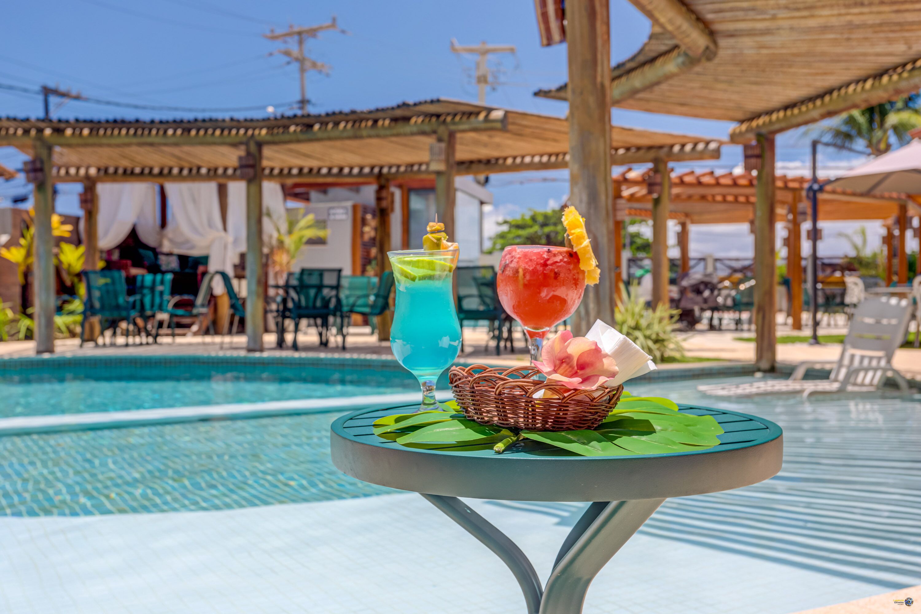 A poolside table with drinks and a basket, surrounded by lounge chairs and a covered seating area.