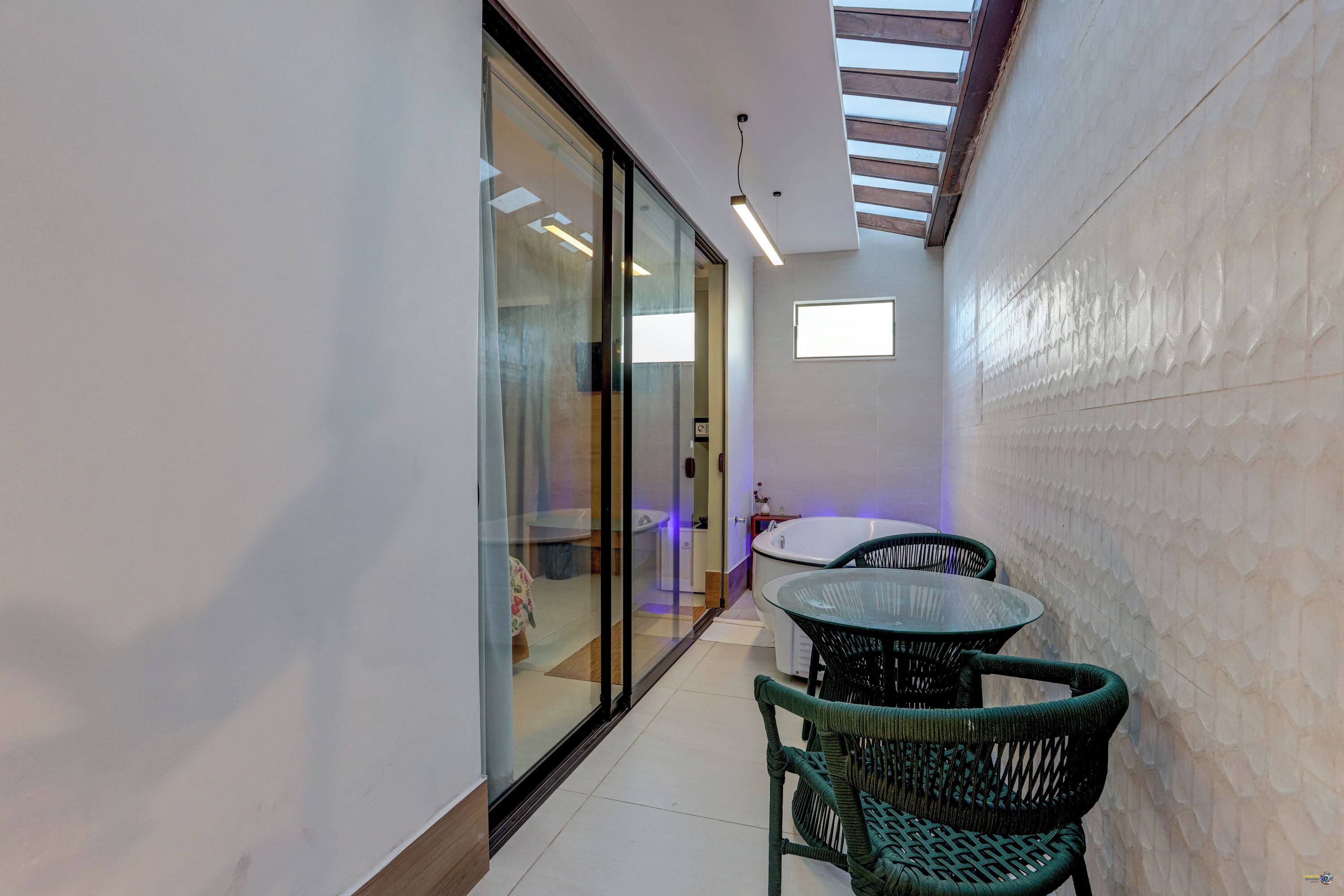 A narrow corridor with a glass door, a round table with chairs, and a bathtub in the background.