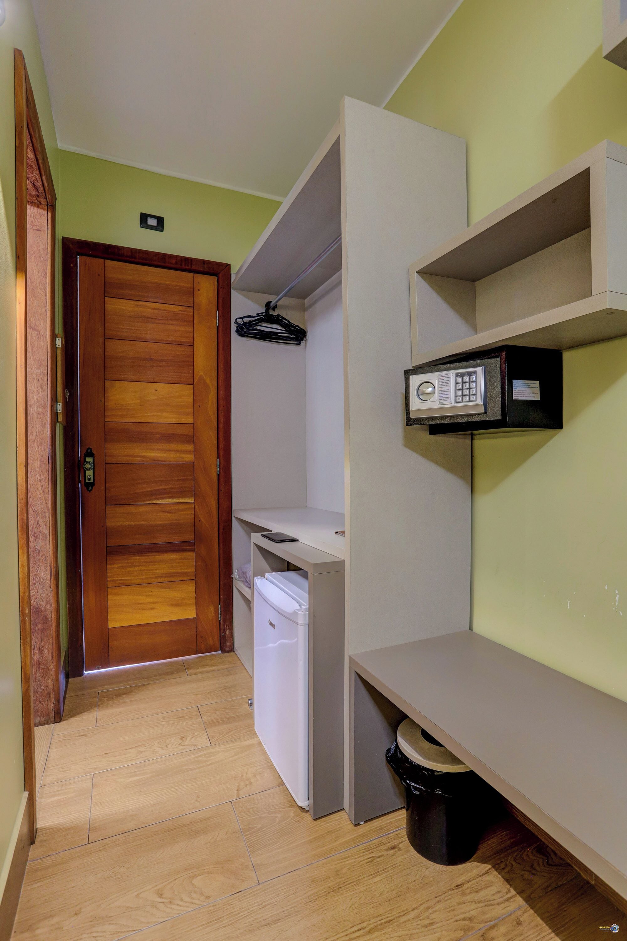 Un pasillo estrecho con una puerta de madera, un cubo de basura blanco, un electrodoméstico pequeño blanco y una caja fuerte sobre una estantería.