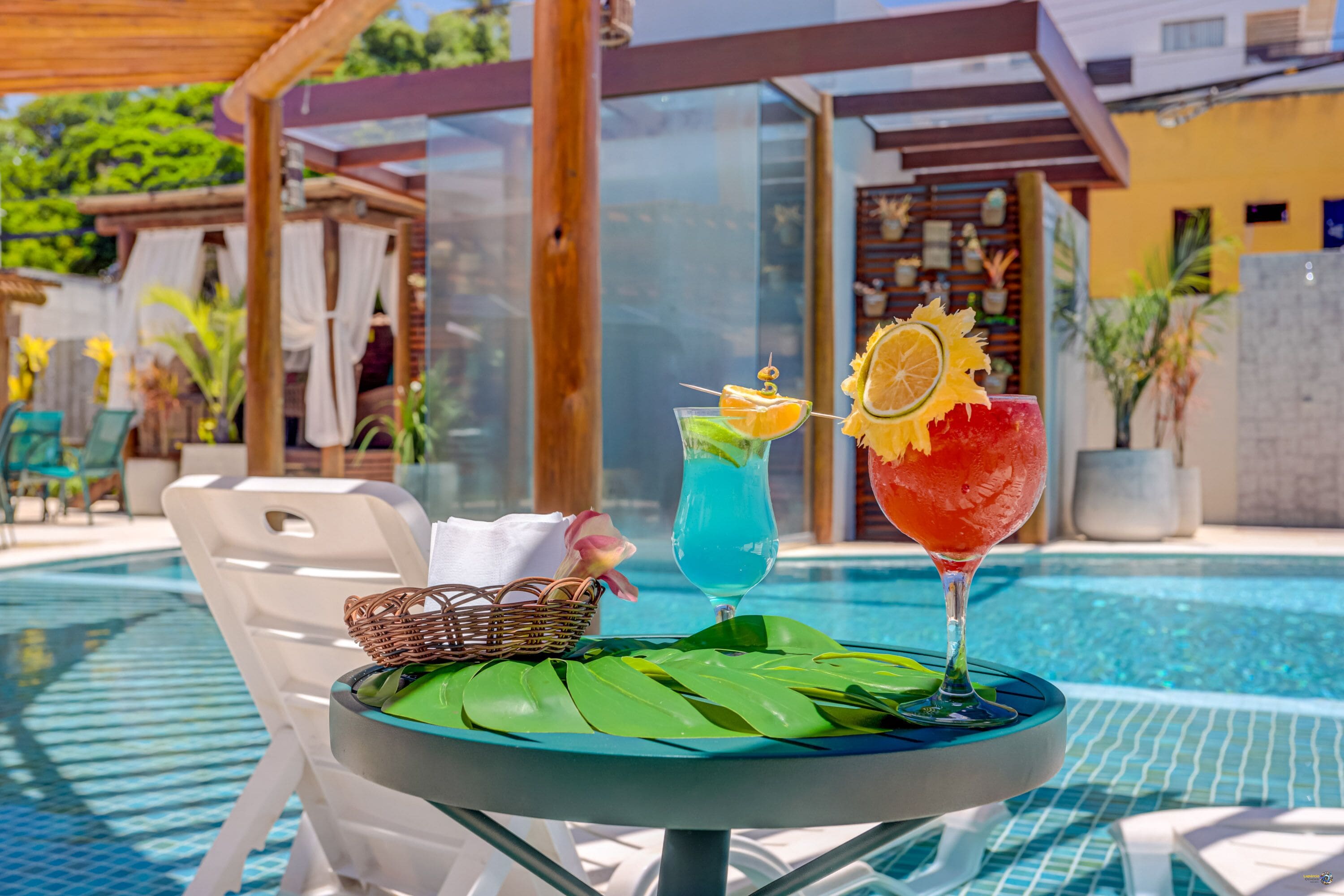 A poolside table with two drinks, a wicker basket, and a green leaf.