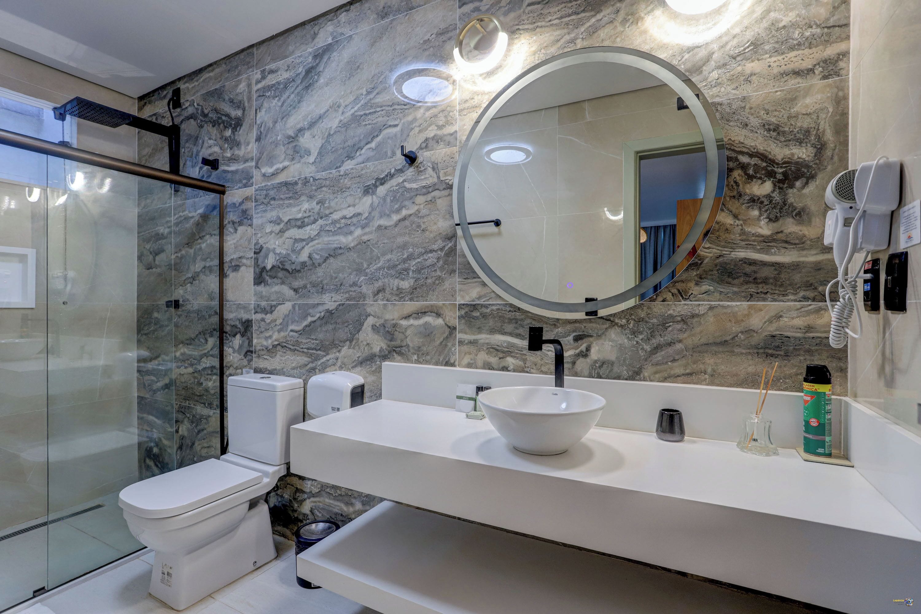 A modern bathroom with a glass-enclosed shower, a white toilet, a white sink with a bowl, a large mirror, and a hairdryer mounted on the wall.