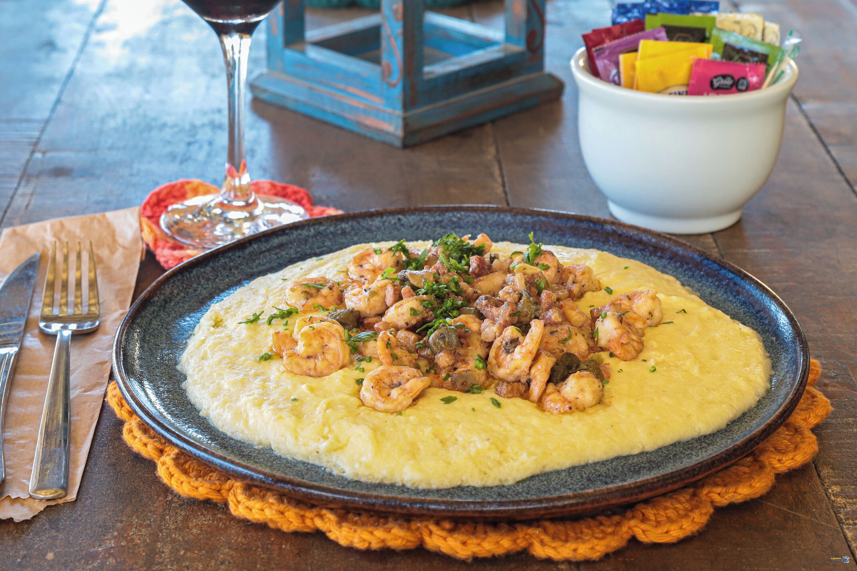 Un plato de camarones y polenta sobre una mesa, junto a una copa de vino y un bol de aperitivos.