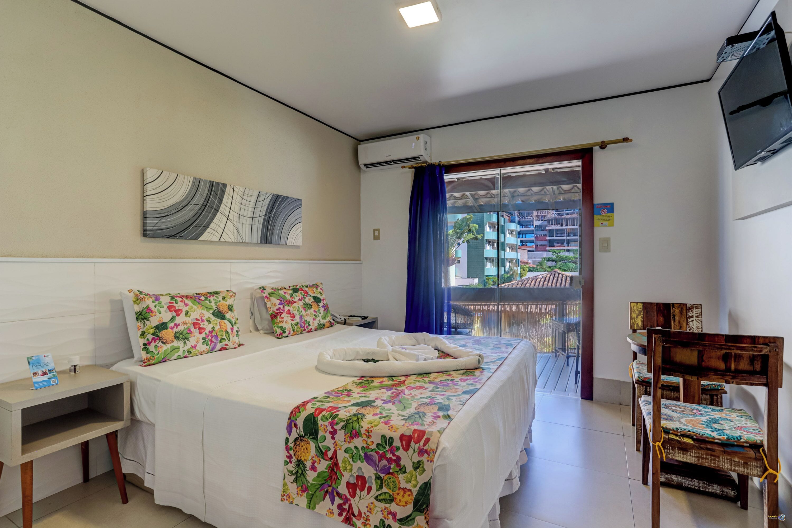A hotel room with a bed, a bedside table, a chair, a TV, and a balcony with a view of buildings.