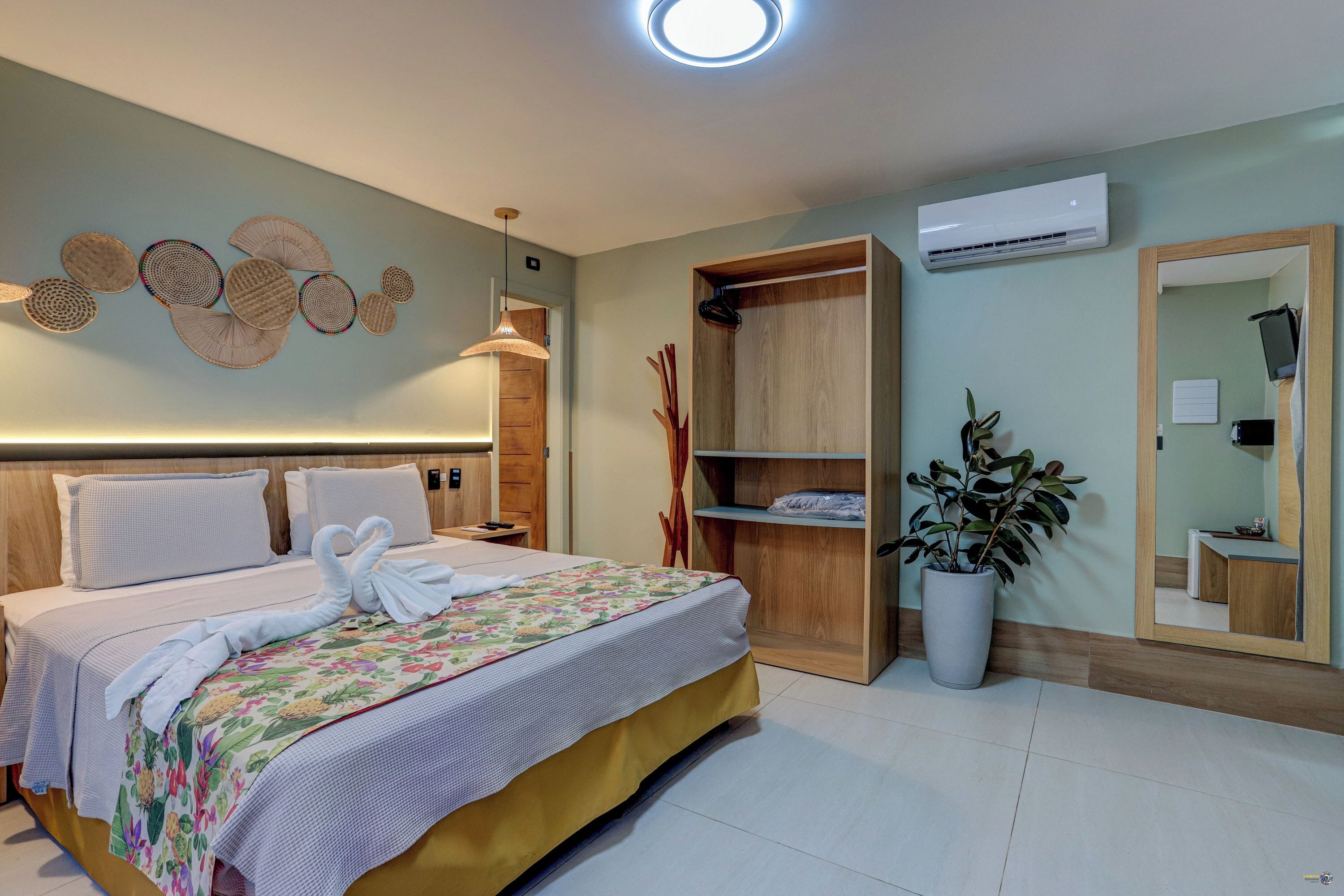 Habitación de hotel con cama, armario de madera, planta en maceta, espejo y equipo de aire acondicionado.