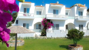 Un edificio blanco con balcones, una sombrilla de paja y un jardín con flores rosadas.