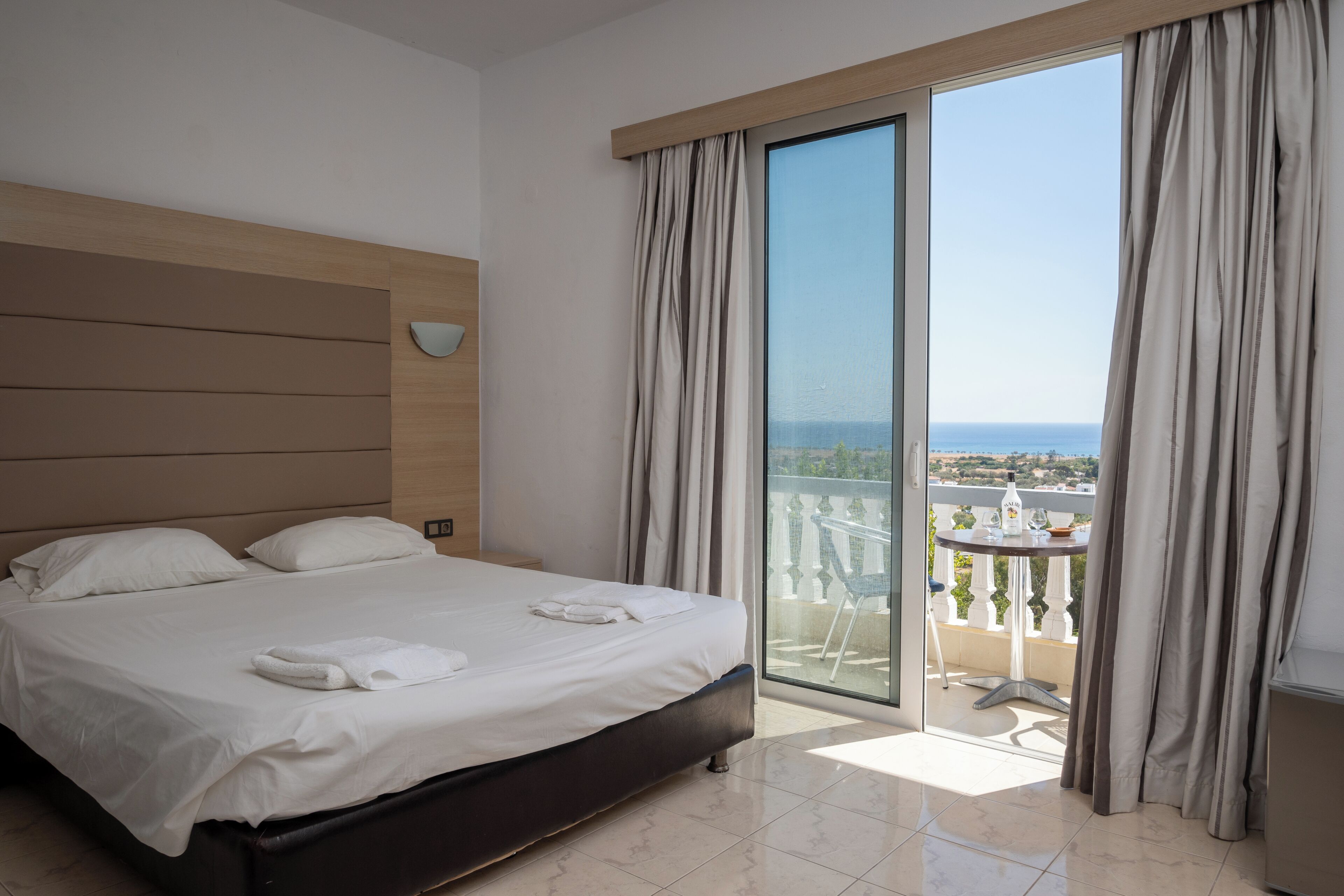 A hotel room with a large bed, a balcony with a table and chairs, and a view of the sea.