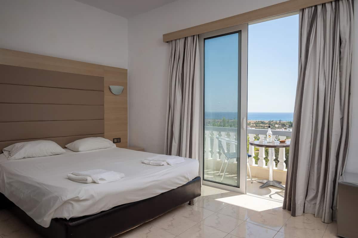 Una habitación de hotel con una cama grande, un balcón con mesa y sillas, y vista al mar.