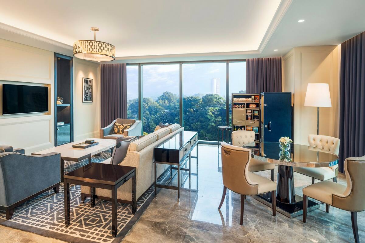 A modern hotel room with a large window, a sofa, a dining table, and a minibar.