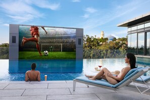 A poolside area with a large screen displaying a soccer game, a lounge chair, and a person sitting by the pool. - The St. Regis Kuala Lumpur (Kuala Lumpur)