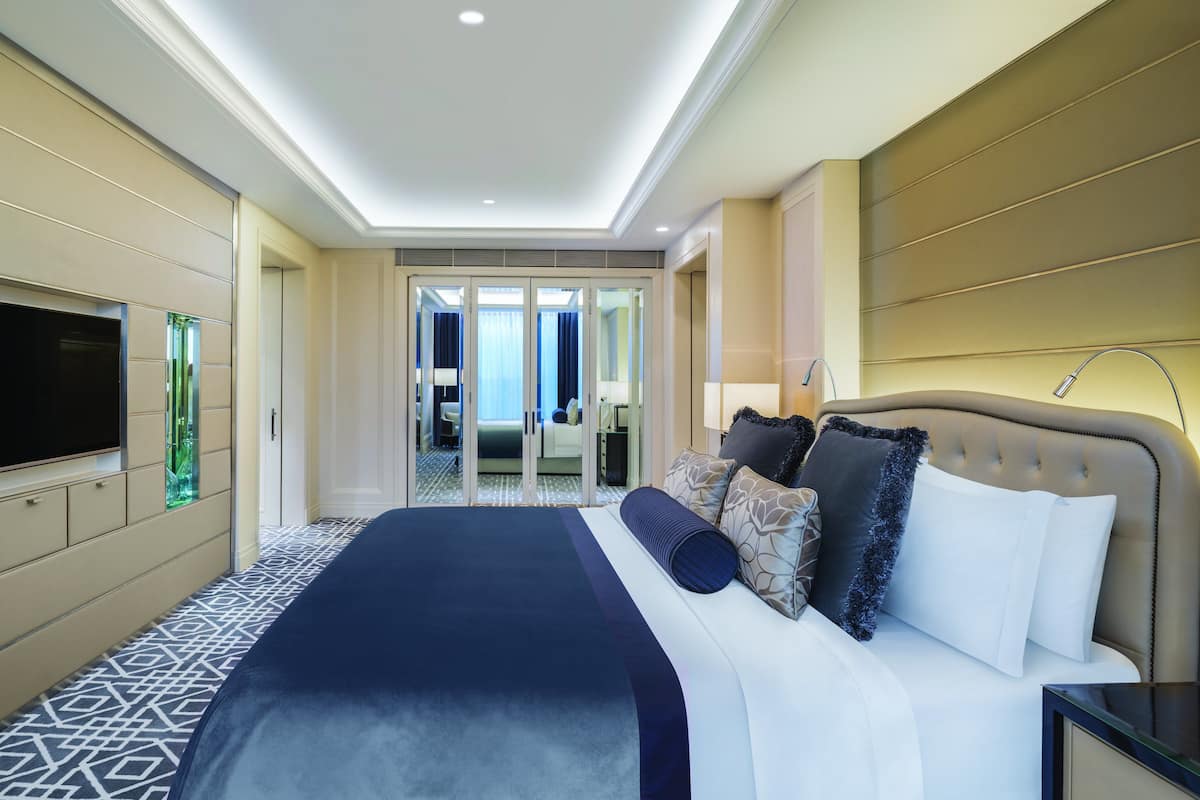 A modern hotel room with a large bed, a flat-screen TV, a minibar, and a bathroom visible through a glass door.