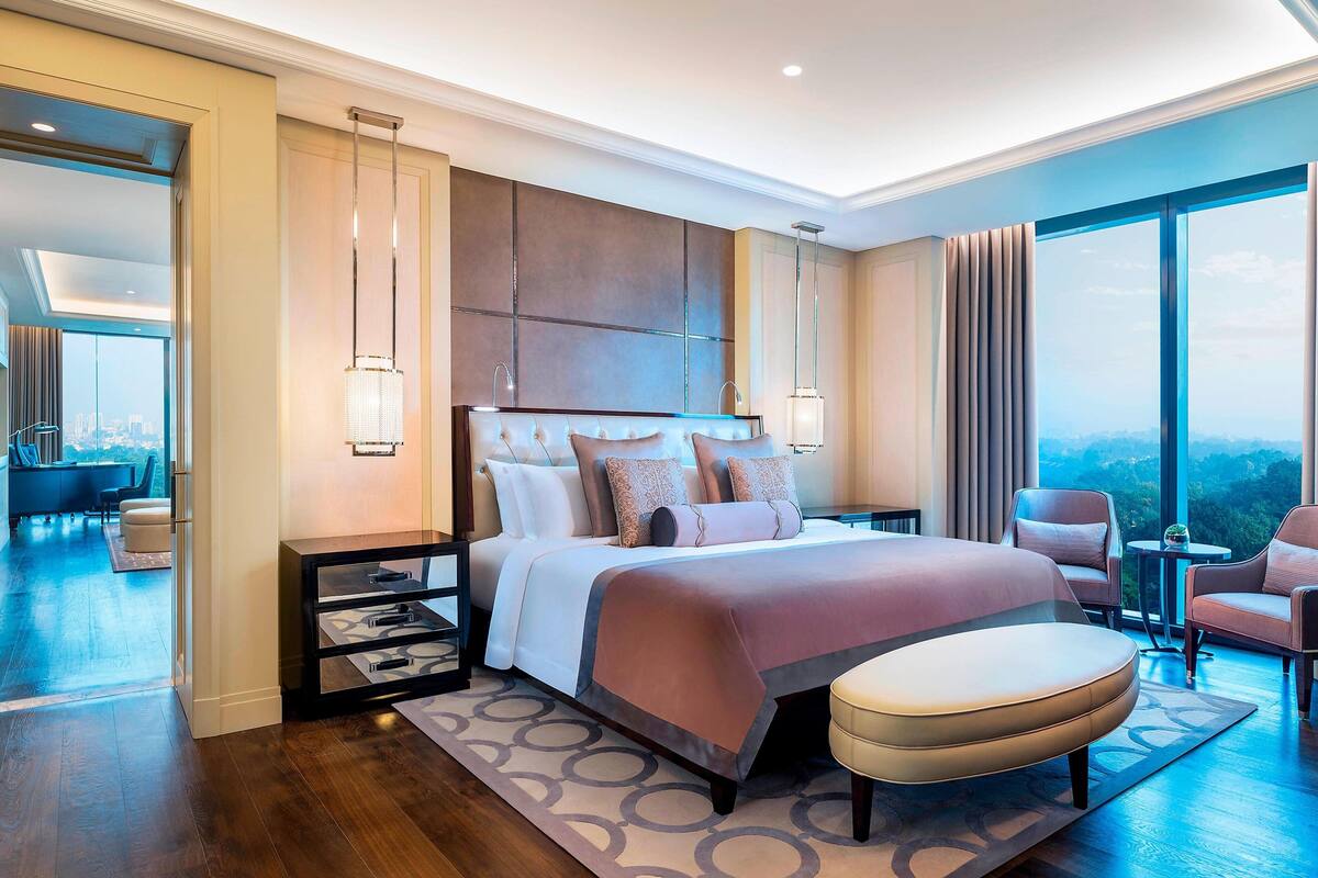 A modern hotel bedroom with a large bed, a bedside table, a chair, and a view of the city.