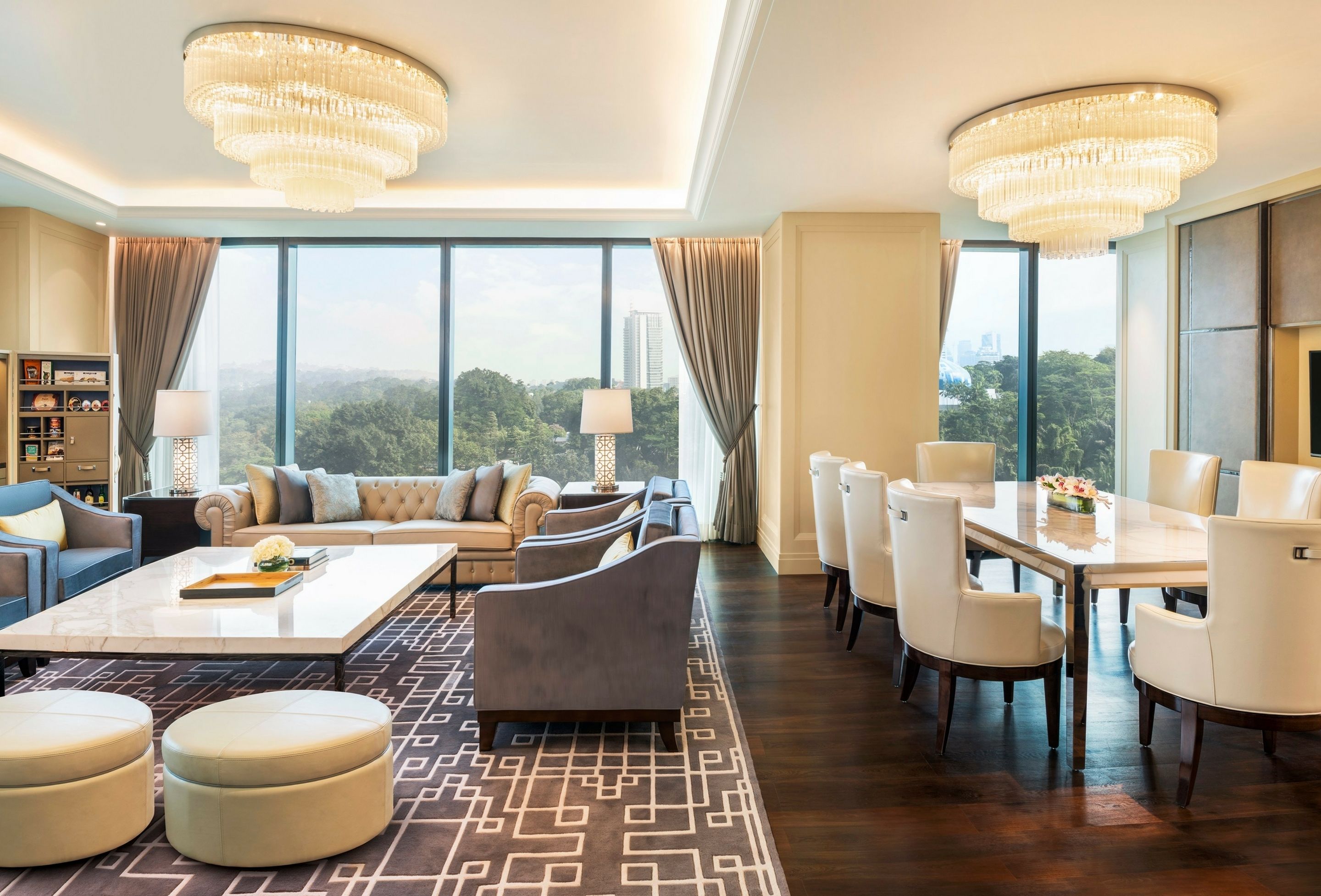 A spacious hotel room with a large window offering a city view, a chandelier, a sofa set, a dining area, and a bookshelf.