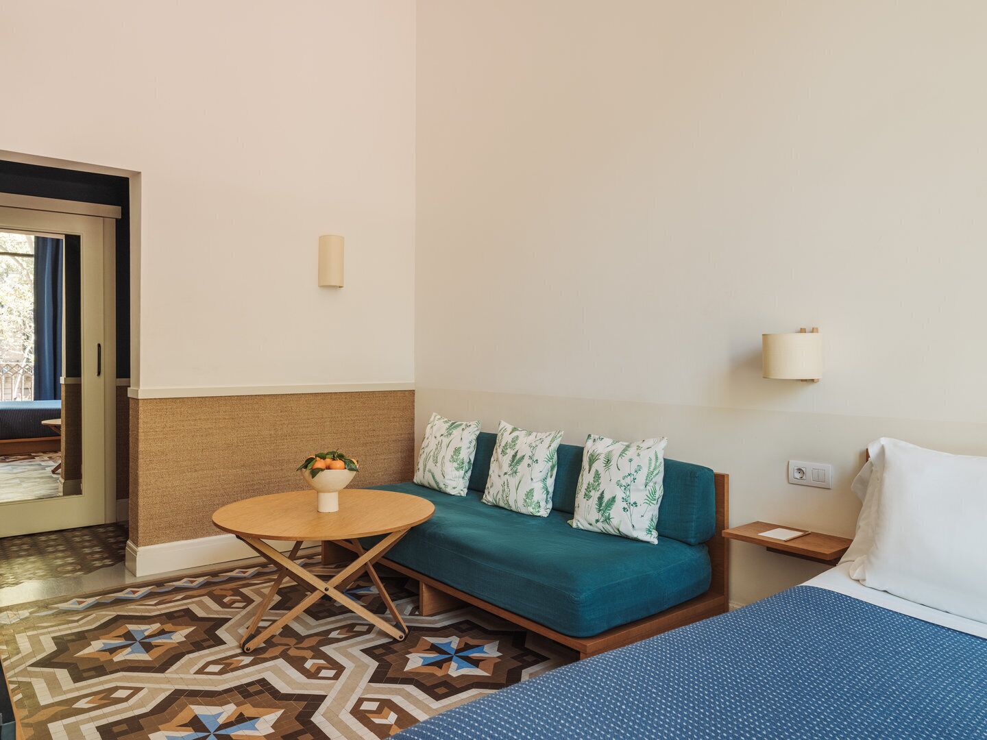 Una habitación con un sofá, una pequeña mesa redonda con un frutero, piso con diseño y una cama con edredón azul.