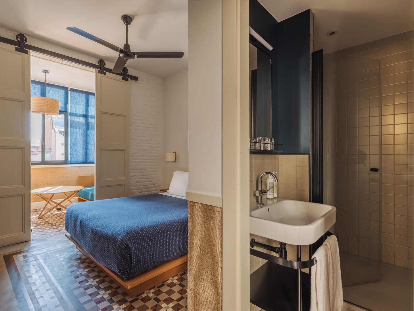A bedroom with a bed, a ceiling fan, a window with a view, a small table, and a patterned floor.