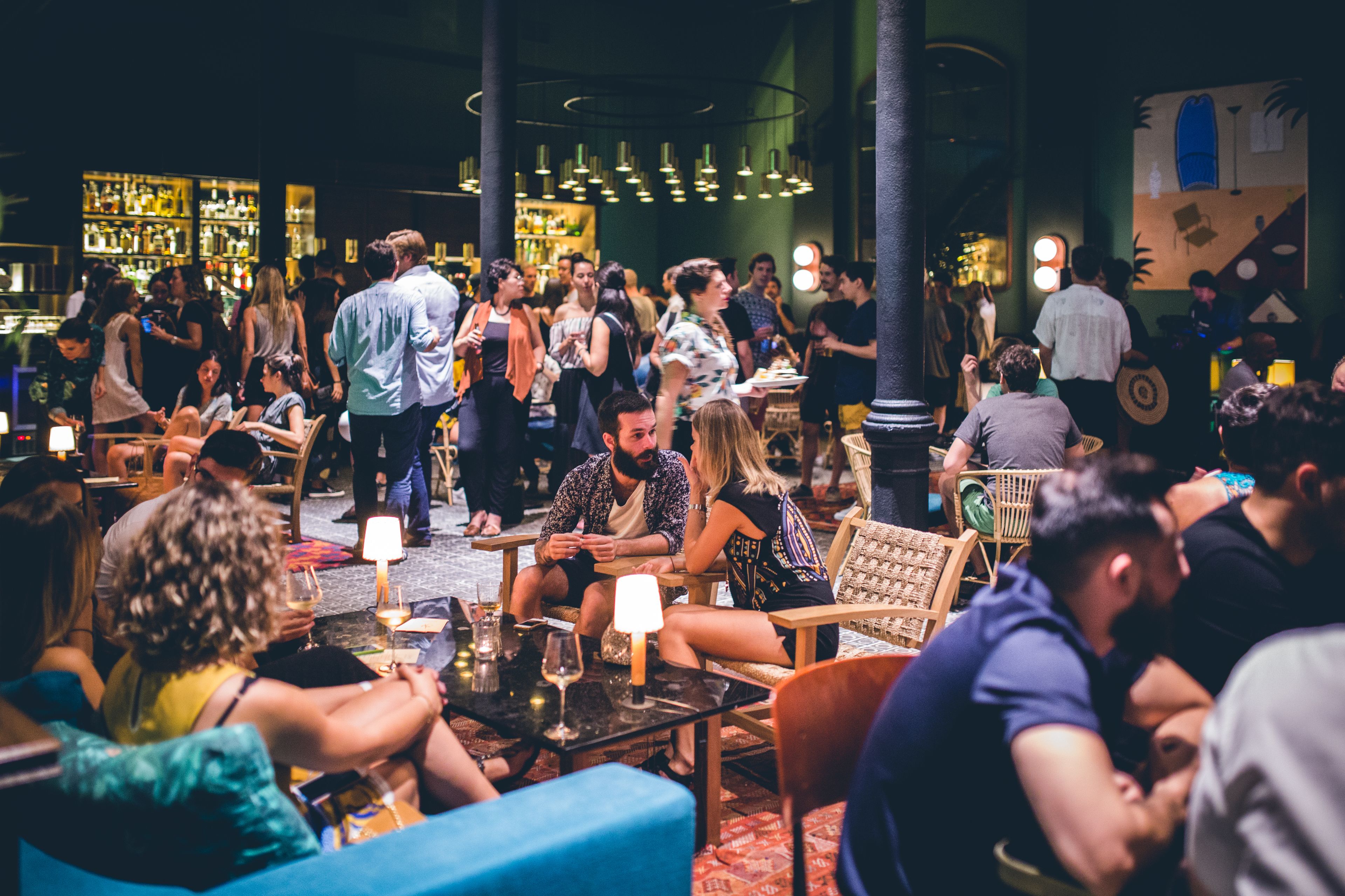 Un espacio social interior concurrido, con mesas y sillas, una zona de bar al fondo y un mural en la pared.