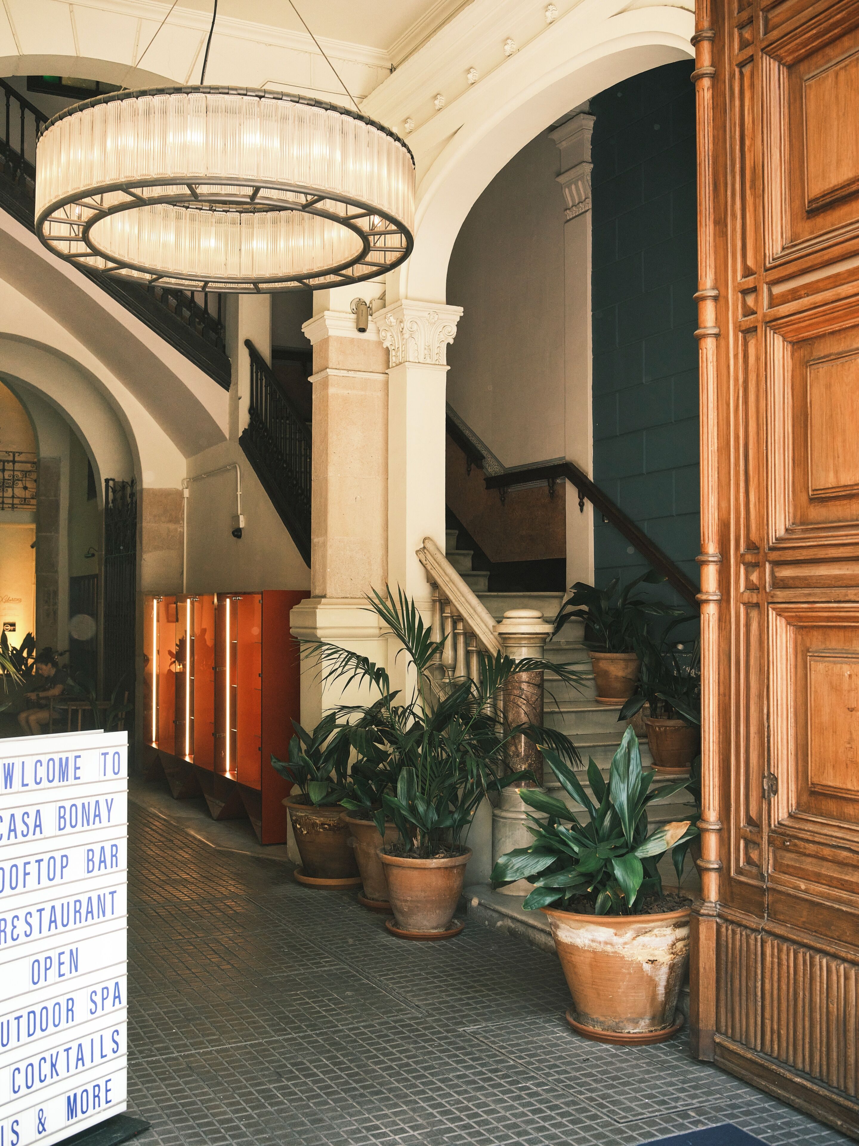 Um hall de hotel com um lustre imponente, uma escadaria e plantas em vasos.