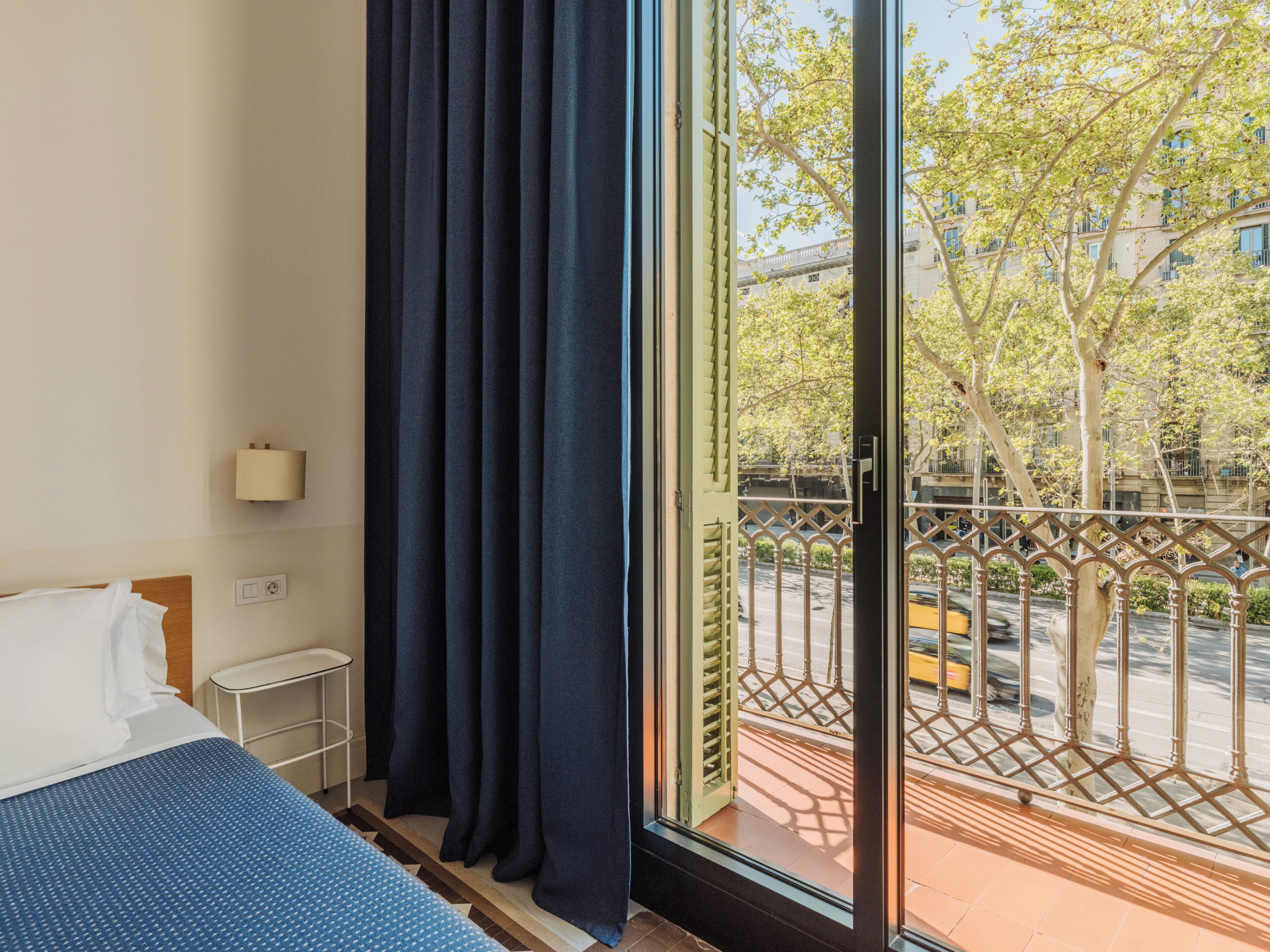 Une chambre d’hôtel avec un lit, une table de chevet, une lampe, une fenêtre avec des rideaux et une vue sur la rue et les arbres.