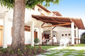 Un'area salotto esterna coperta con un pergolato in legno, un albero grande e un giardino curato con lastre di pietra.