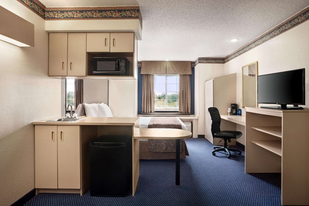 Quarto de hotel compacto com cozinha compacta, cama, escrivaninha com cadeira, televisão e janela com cortinas.