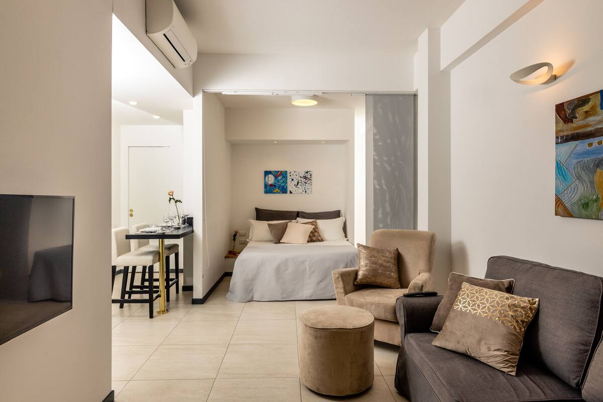 A modern hotel room with a bed, a sofa, a small round ottoman, a desk with a chair, and a TV.
