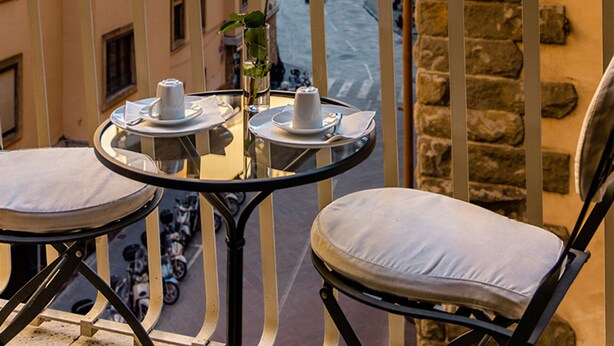 A balcony with a table set for two, a vase with a single yellow rose, and a view of a river and buildings in the background. - MyFlorenceHoliday Gold Bridge (Florence)