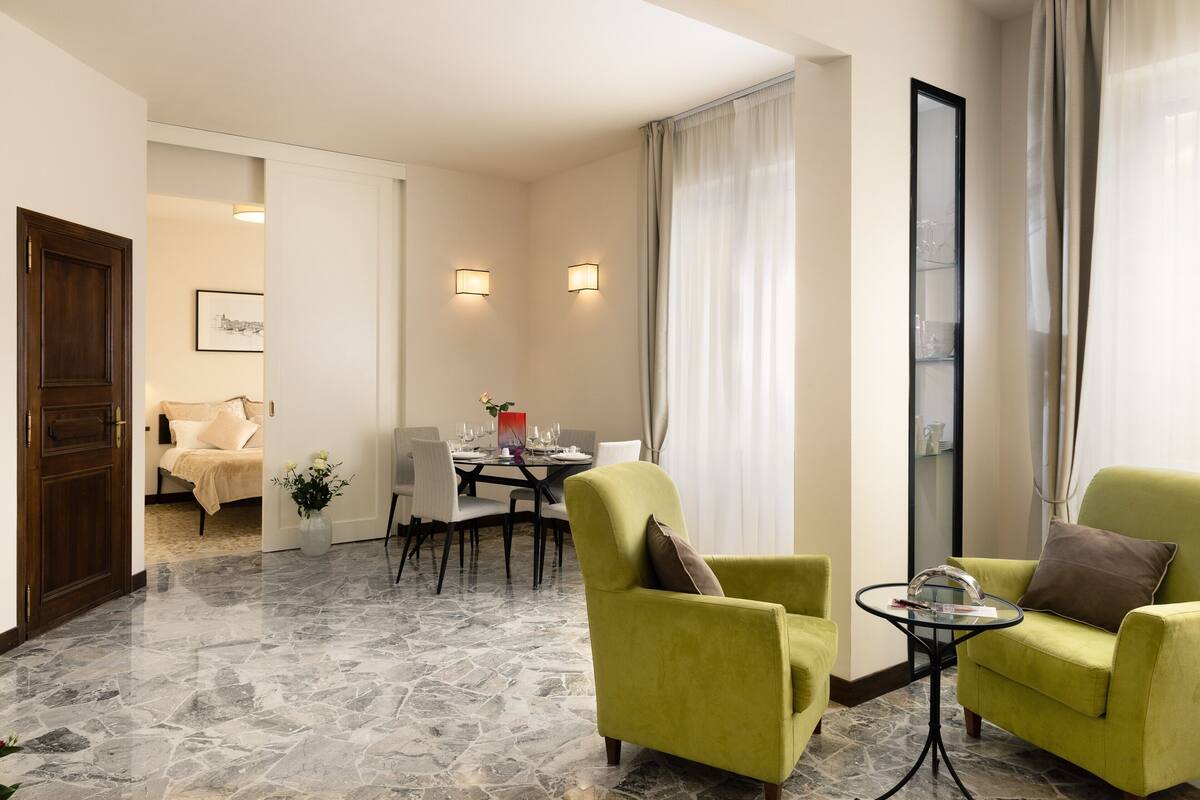 A hotel room with a dining area, a green armchair, a small table, and a large mirror.