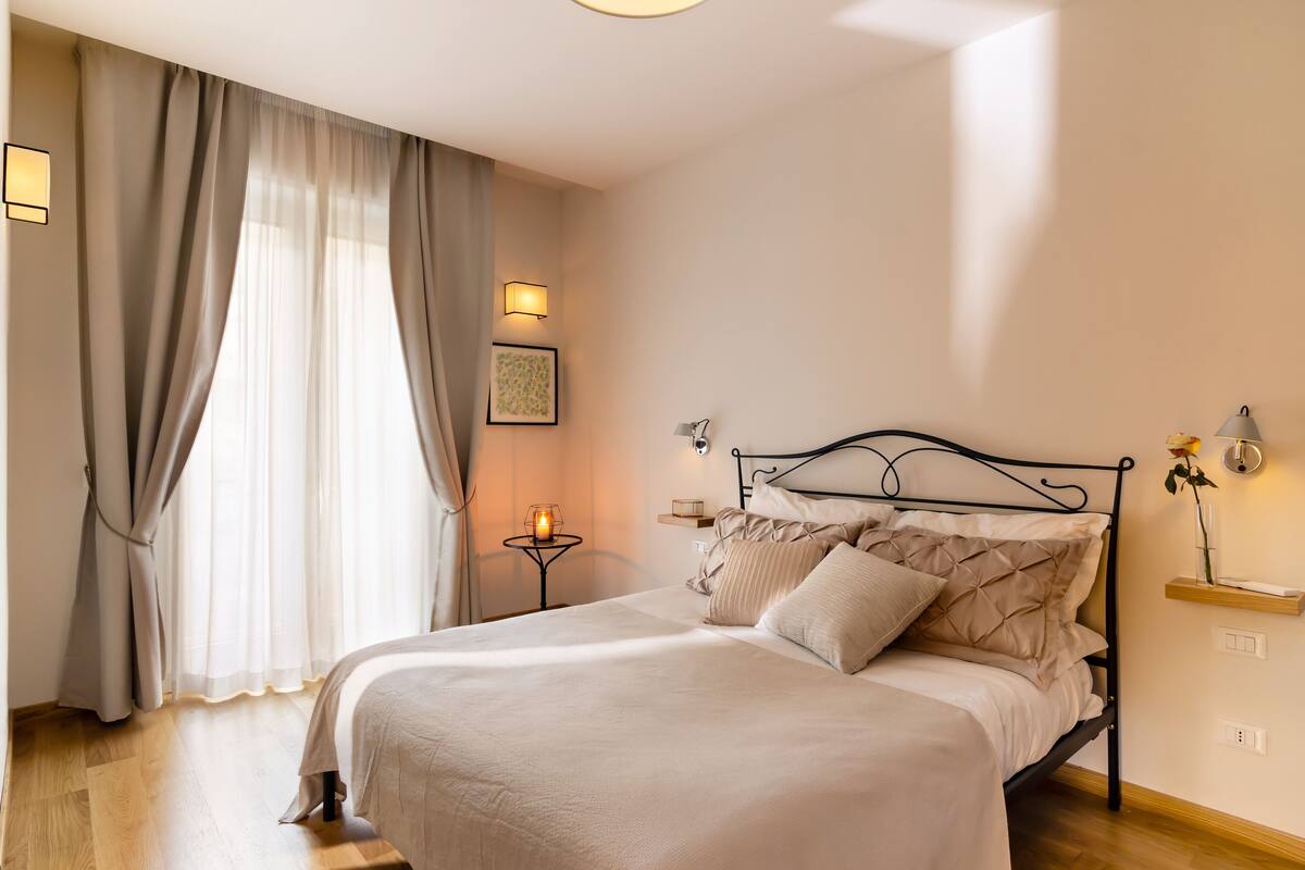 A neatly made bed with a headboard, a bedside table with a lamp, and a wall-mounted shelf with a vase.