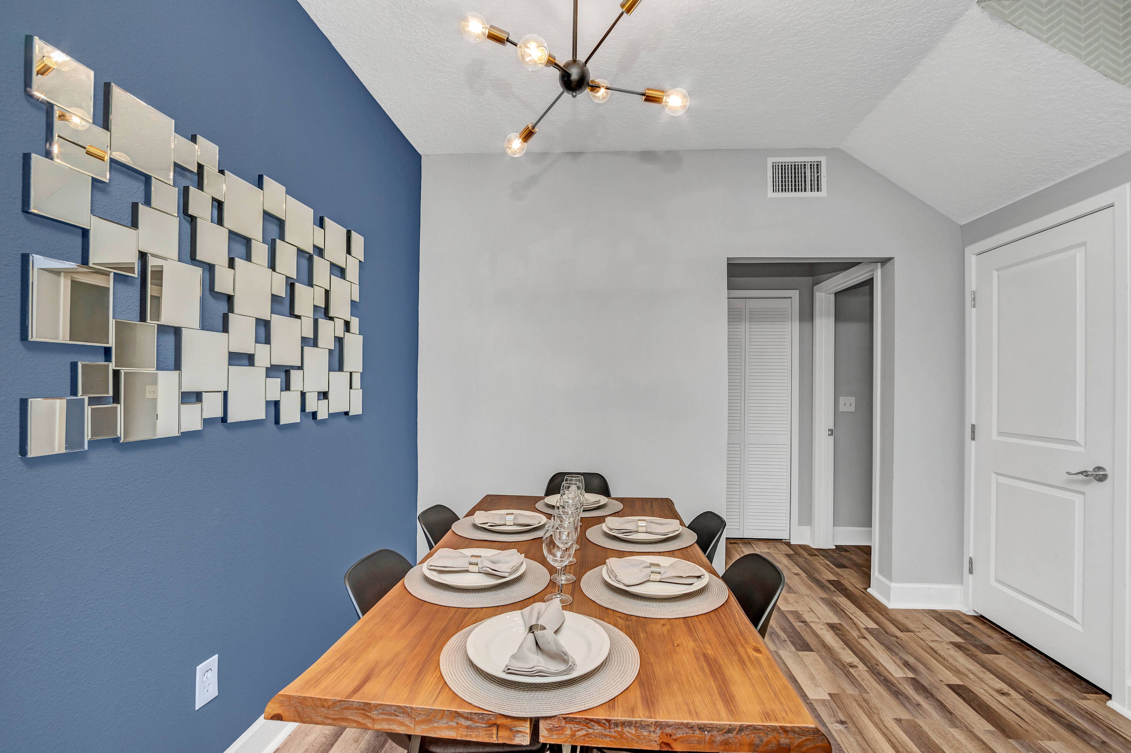 Un comedor con una mesa de madera dispuesta para seis comensales, una instalación artística moderna en la pared y una lámpara de techo.
