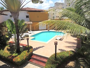 A pool area with a clear blue pool, surrounded by palm trees and a yellow building. - Marinas Pousada (Ilhéus)